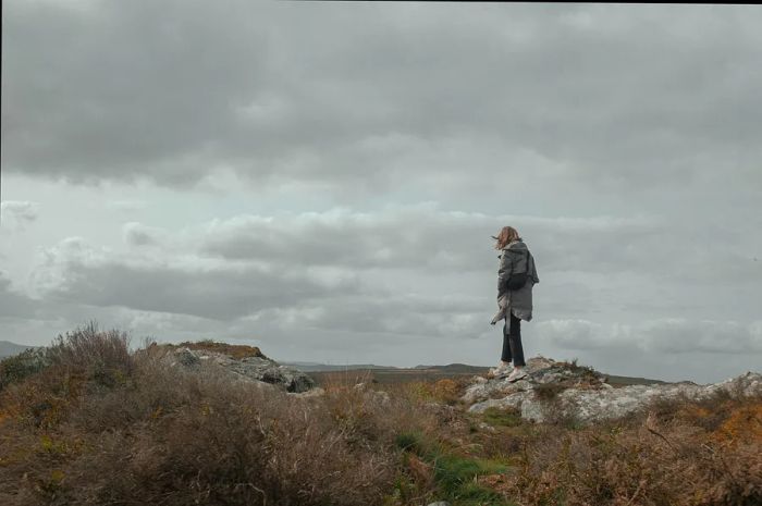 A woman in a rain jacket and hiking boots gazes over the rugged expanse of the Monts d'Arrée