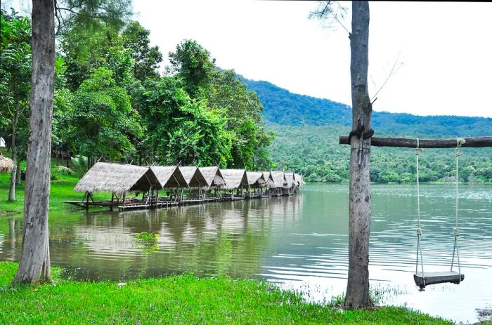 A series of bamboo huts line the edge of Huay Tung Tao, a man-made reservoir near Chiang Mai, with a swing hanging from a nearby tree in the foreground.