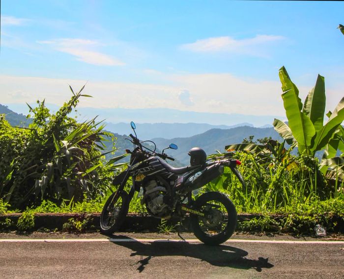 A motorbike rests at a picturesque viewpoint along the Samoeng Loop near Chiang Mai, with a helmet placed on the seat and expansive views of lush green valleys in the background.