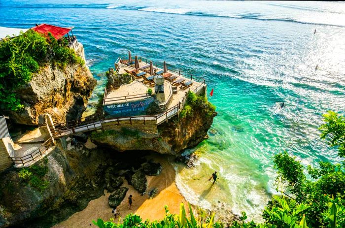 A surfer making their way to the ocean near a steep cliff topped by a bar-lounge.