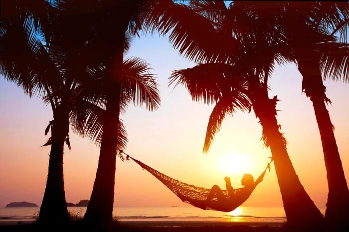 Silhouette of a woman lounging in a hammock on the beach at sunset.