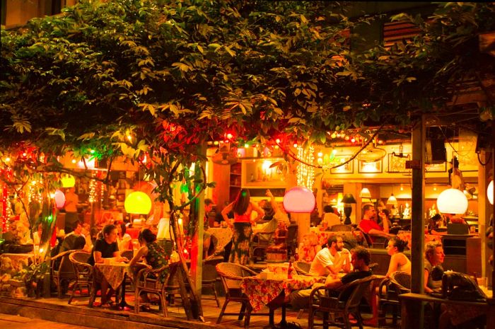 Patrons at the Bangkok Bar in Banglamphu savor their chilled, golden lager under the warm glow of the bar's low-hanging lights, situated near Khao San Road.