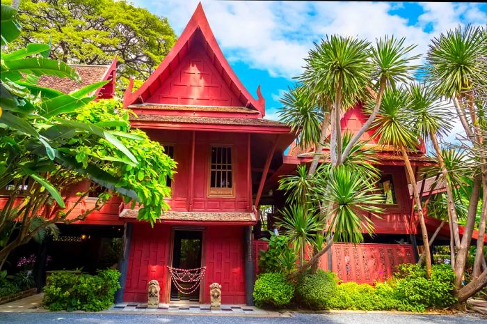 The Jim Thompson Museum features a warm ochre exterior, with the wooden structure slightly obscured by surrounding trees.