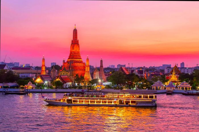 A white river cruise ship glides past the stupa of Wat Arun in Bangkok as the soft glow of an apricot-hued sunset envelops the scene.