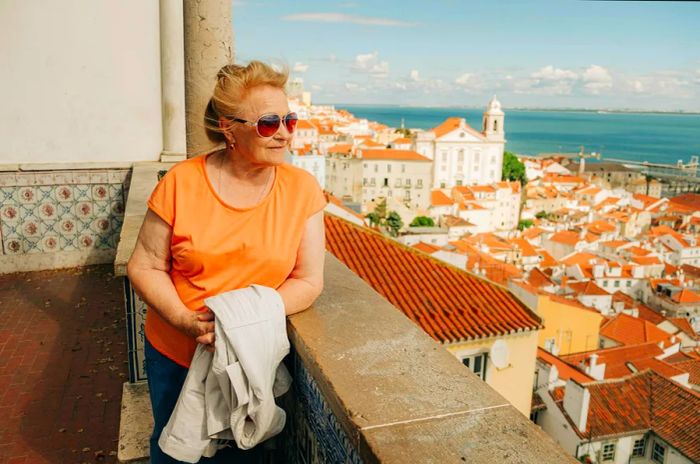 An elderly woman gazes out over the terracotta rooftops of a riverside city from a viewpoint.