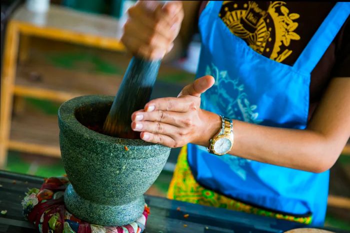 A woman wearing a light blue apron, her head out of view, grinds herbs with a gray pestle and mortar during a cooking class in Bangkok.