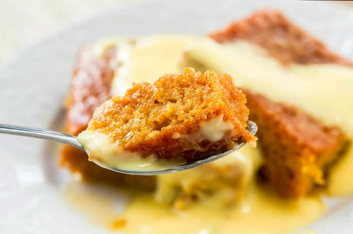 A fork holds a scoop of steaming malva pudding, generously covered in yellow custard.