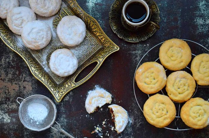 Sesame shortbread kahk cookies filled with dates from Egypt. Some cookies are presented on a decorative gold tray dusted with icing sugar, with one broken in half to reveal the filling, alongside a cooling rack and a small cup of tea.