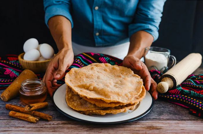 A man showcases the flat, disc-shaped Buñuelos dessert on a table adorned with traditional Mexican patterns.