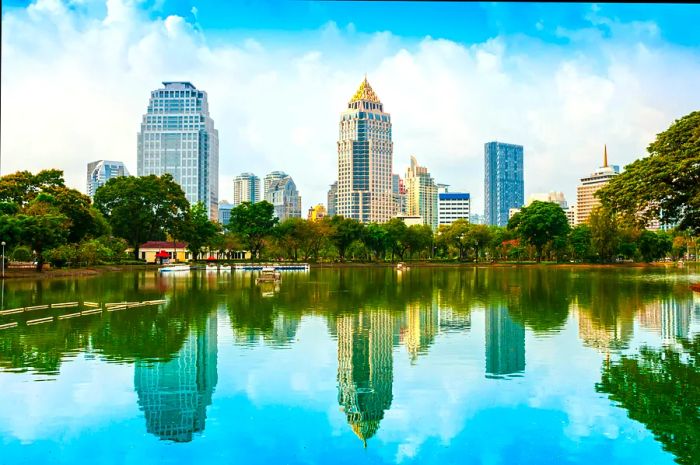 The towering skyscrapers of Bangkok’s skyline rise against a vibrant blue sky above the serene, shimmering lake within Lumphini Park.