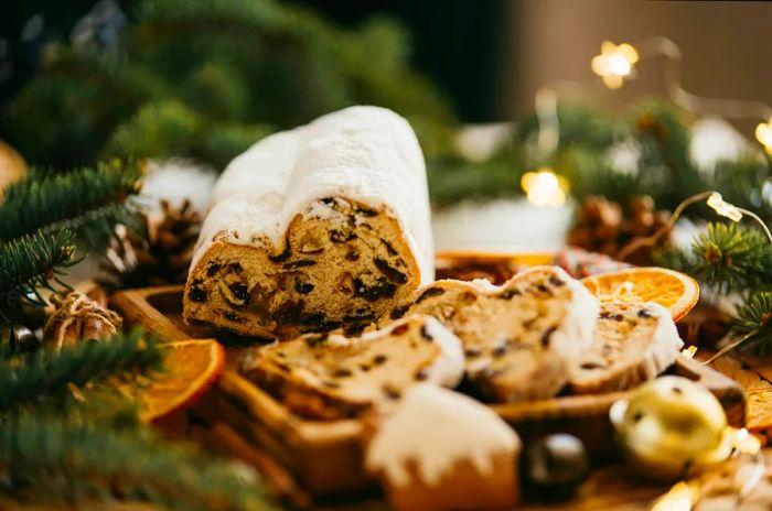 Slices of traditional Christmas stollen are displayed on a wooden surface. This German dessert features a cake with nuts, raisins, marzipan, and dried fruits, beautifully arranged on a cutting board for holiday baking.