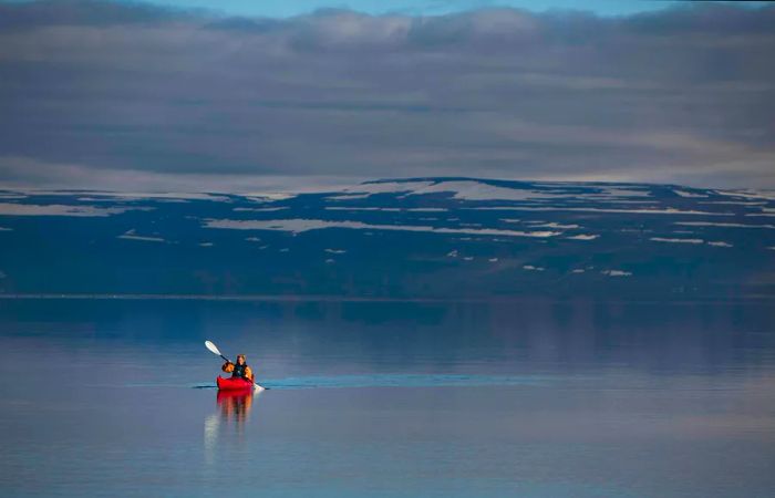 A woman in a red kayak glides across a serene lake with snow-capped mountains in the background.