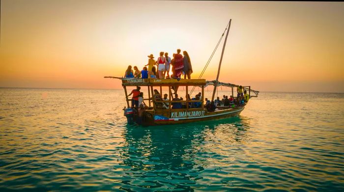 A traditional Dhow filled with tourists, both inside and on the roof, glides on the water at sunset in Zanzibar