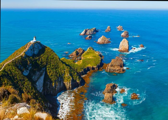 Nugget Point Lighthouse, New Zealand, perched atop a forested coastal peak, with a series of rocky islands extending into the sea beyond.