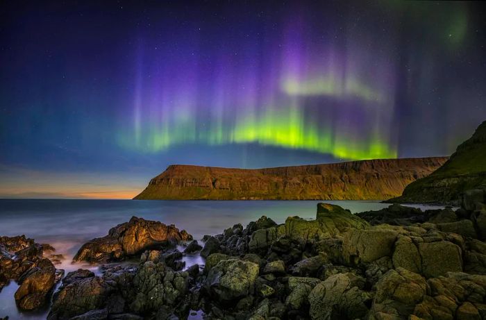 The night sky dances with purple, green, and yellow as the Northern Lights illuminate the cliffside.