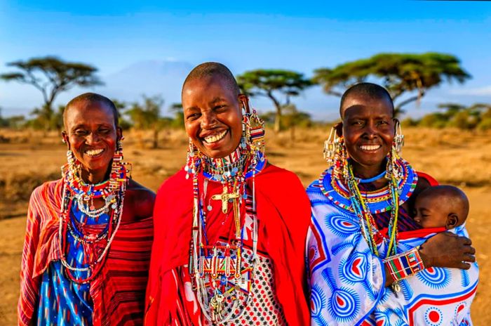 Three smiling Maasai women dressed in vibrant prints, with one holding a small baby.