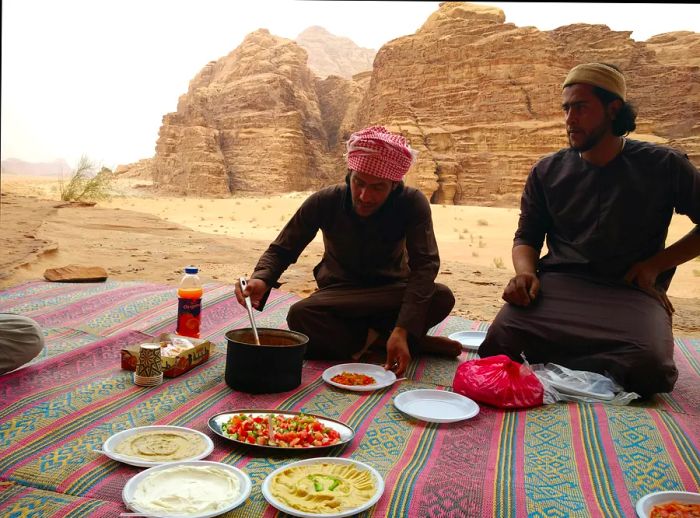 Two Bedouin men share a meal on vibrant, striped carpets in the Jordanian desert.