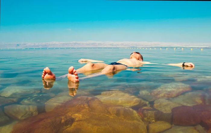 A man wearing sunglasses and a dark Speedo floats on his back in the salty blue waters of the Dead Sea, Israel