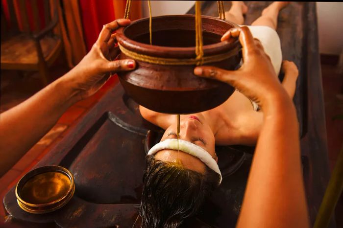 A woman relaxes on her back in a spa, as a golden liquid is gently drizzled onto her forehead during an Ayurvedic wellness session.