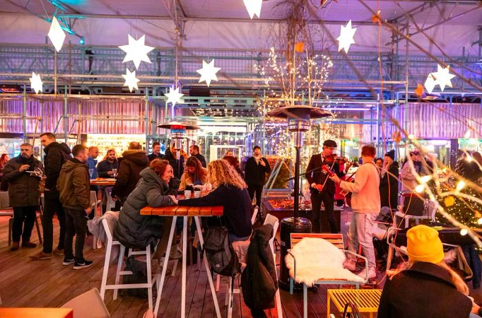 A crowd bundled in winter coats and hats sits at tables in Prague's Manifesto Market Anděl, with a band playing nearby. The outdoor market is adorned with star-shaped lights.