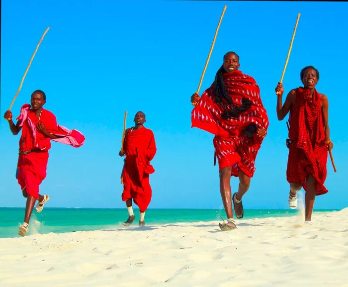 Four Maasai warriors dressed in red, holding sticks, leaping on a beach in Zanzibar