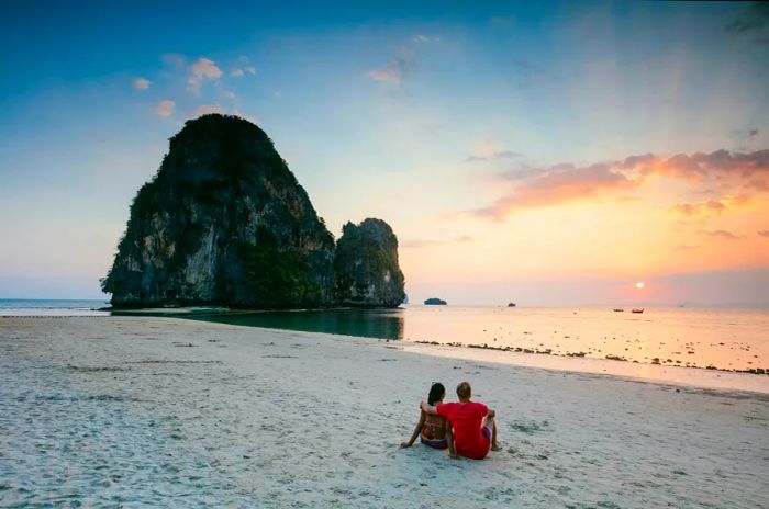 A couple enjoys the sunset on a sandy beach, gazing out at the sea where the sun dips behind a striking rocky formation.