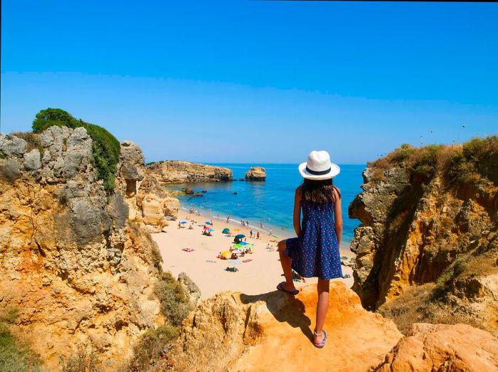 A young girl peers over a cliff, looking down at a sandy beach bustling with families enjoying the sunny coast.