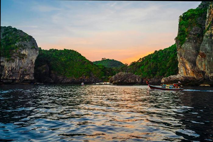 A view from the water towards the shore, featuring large rocky cliffs adorned with greenery. A small, vibrant fishing boat passes by.