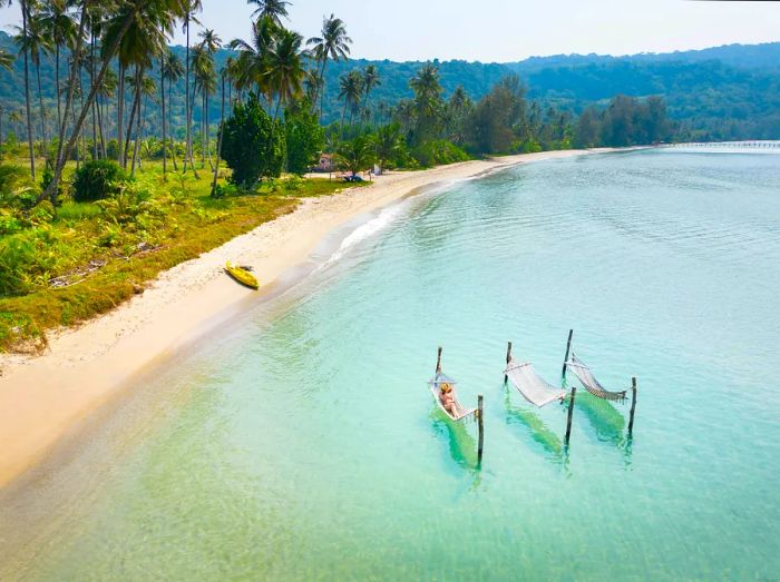 A woman relaxes in a hammock suspended over the sea, with an empty sandy beach lined with palm trees stretching into the distance.