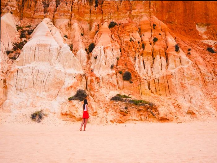 A woman strolls along the sandy shore in front of a towering cliff at Praia da Falésia beach, located in the Algarve, southern Portugal, Europe.