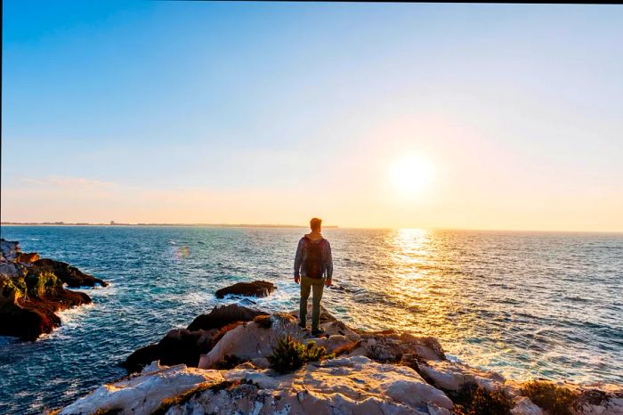 A young man stands on a cliff, gazing at the sunset over the ocean at Praia Baleal, Portugal, Europe.