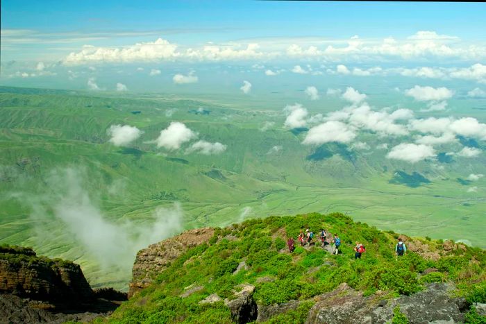 A group descends from the summit of Ol Doinyo Lengai into the lush Rift Valley. The escarpment of the Rift Valley looms in the background, while volcanic smoke billows from a crack in the foreground.