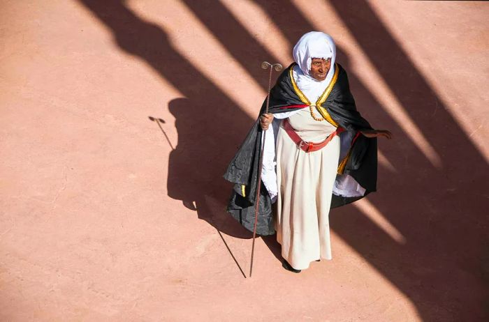 An elderly African Christian woman in traditional attire casts a shadow in the sunlight.