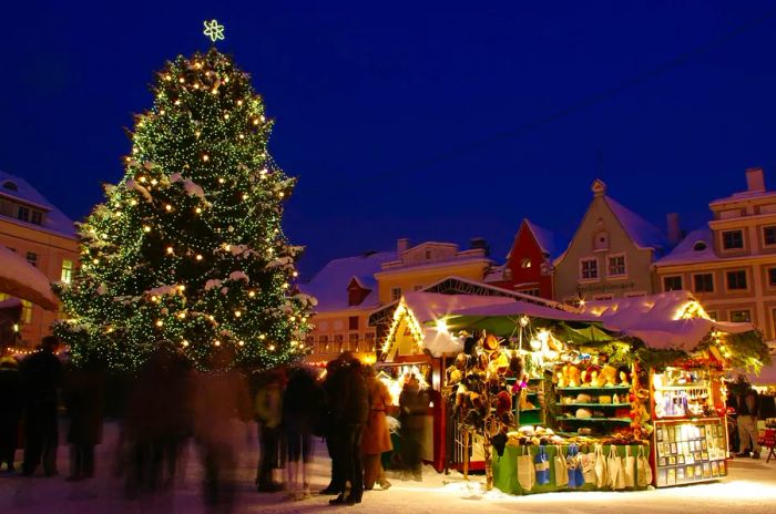 A nighttime Christmas market in Tallinn, Estonia, showcases a large, decorated Christmas tree surrounded by wooden stalls offering festive goods, with blurred figures wandering through the market.