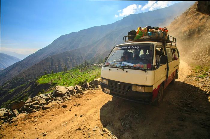 An old, small bus navigates a rugged dirt road in the Peruvian Andes, with a steep drop-off beside it leading into a lush green valley.