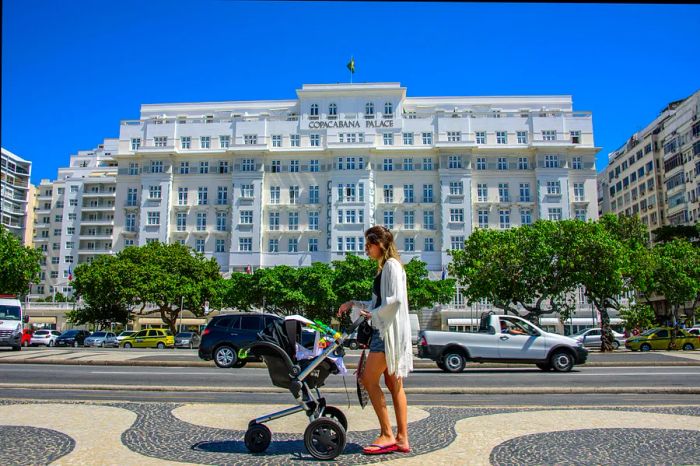 A woman dressed in a white gown and flip flops strolls down Avenida Atlantica with a black stroller, with the iconic Copacabana Palace in the backdrop.