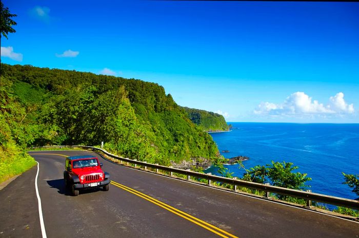 A red car navigates a winding cliffside road alongside the ocean.