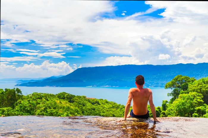 A young man without a shirt gazes at the stunning view of lush hills, clear blue waters, and distant gray, cloud-covered mountains.