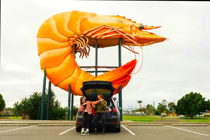 Two kids lean against the trunk of a car parked beneath a massive prawn sculpture.