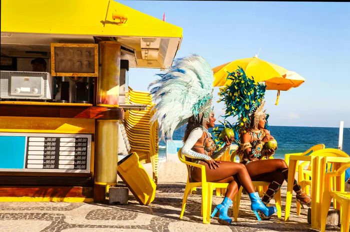 Samba dancers in vibrant costumes with feathers, enjoying coconuts outside a snack stand at Ipanema Beach, with the ocean in the backdrop