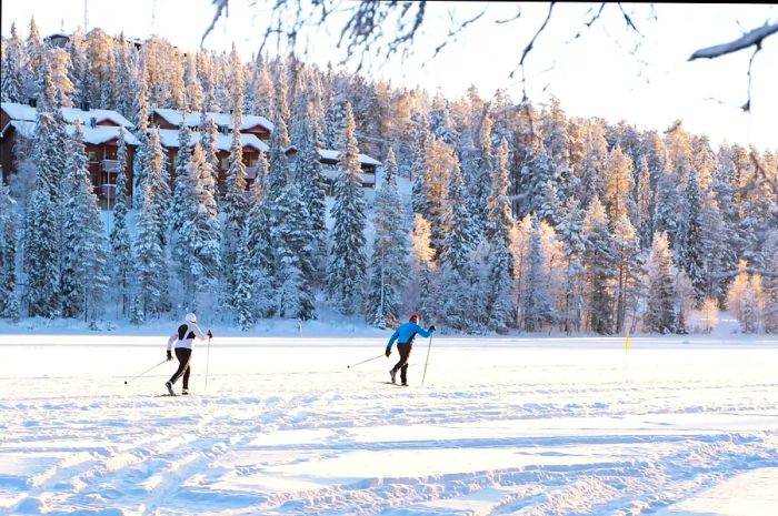 Two athletes cross-country skiing on a frozen lake surrounded by a snow-covered forest.