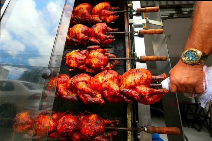 A street restaurant in Salvador, Brazil, showcasing whole chickens on skewers being grilled, with someone turning the skewers using a towel.