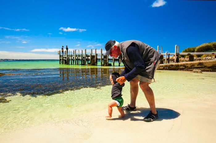 A grandfather guides a toddler's hands as they venture onto the beach, heading straight for the ocean