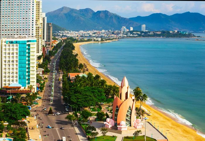 An aerial perspective of Nha Trang reveals a skyline of high-rise buildings on the left and a golden beach stretching alongside the city on the right.