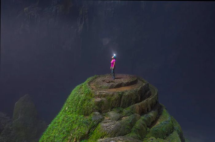 A woman in a helmet with a light stands atop a jagged rock, looking up at an opening in the ceiling of the vast Hang Son Doong cave.