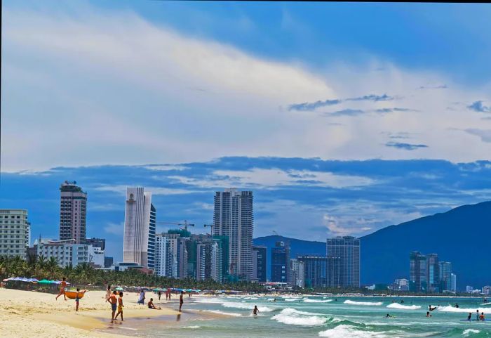 A view showcasing seafront high-rise hotels adjacent to sandy My Khe beach in Da Nang, Vietnam. Numerous people enjoy the beach or swim in the sea.