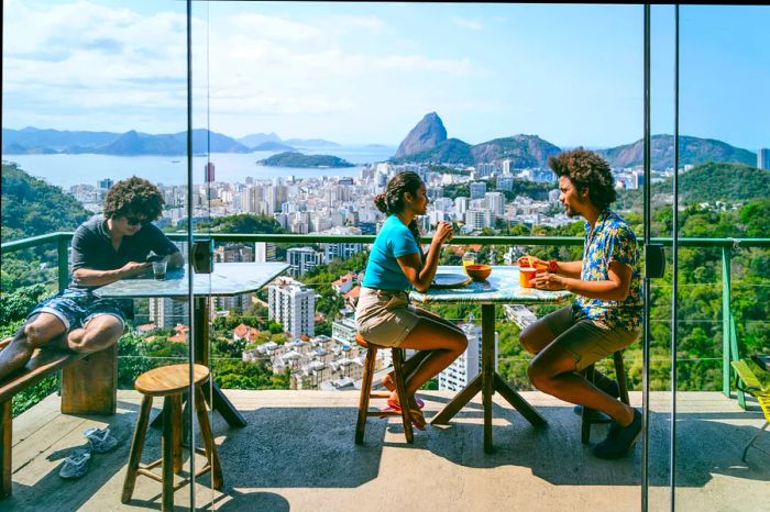 Two individuals converse on a balcony with a view of Rio de Janeiro.