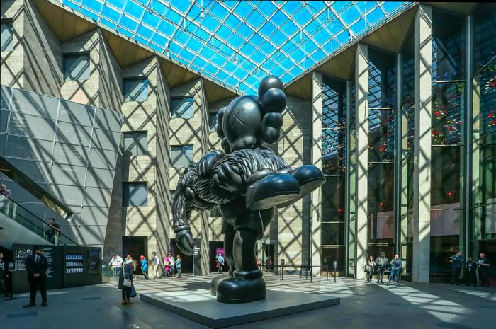 A large black surrealist statue takes center stage in a spacious courtyard beneath a glass ceiling, part of the National Gallery of Victoria in Melbourne.