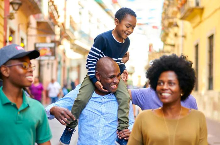 A Black family strolls through Mexico City, with the father lifting the younger son onto his shoulders while the mother and teenage son walk ahead, all smiling.