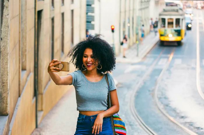 A woman beams as she snaps a selfie in Lisbon, Portugal, with a yellow tram descending the hill in the background.